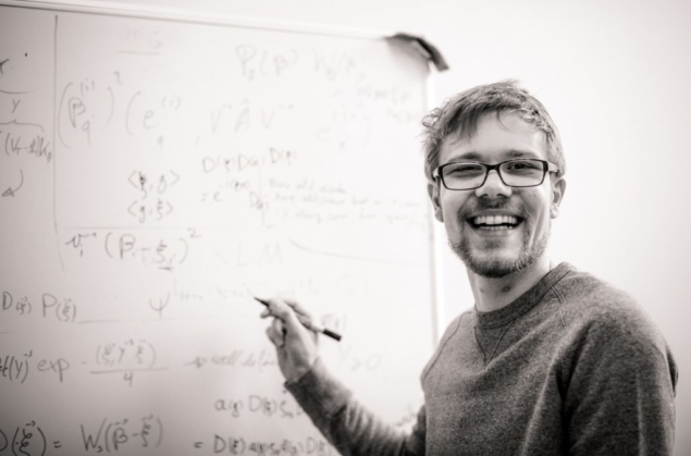 Photo of a smiling Mattia Walschaers standing in front of a whiteboard filled with equations and diagrams